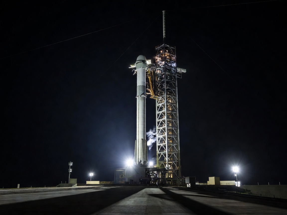A rocket standing on its launch pad at night, lit by floodlights against a dark sky